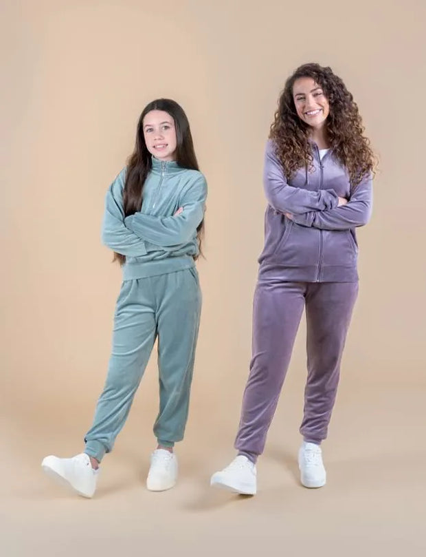 Two young women modeling matching two-piece tracksuits in different colors, standing against a solid beige studio background. The woman on the left wears a sage green set, and the woman on the right wears a lavender-purple set.