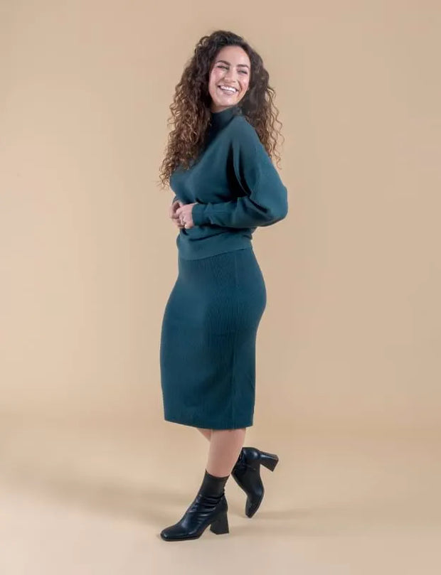 A woman modeling a dark teal ribbed knit mock-neck sweater and matching midi skirt set, paired with black ankle boots, against a solid beige studio background.