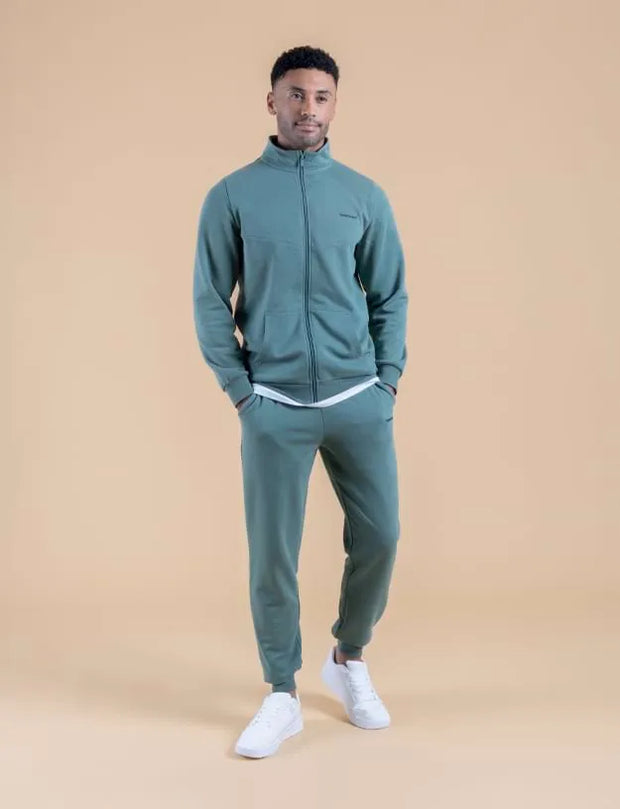 A man modeling a matching two-piece tracksuit in a muted teal green, featuring a zip-up jacket and jogger pants, styled with white sneakers against a solid beige studio background.