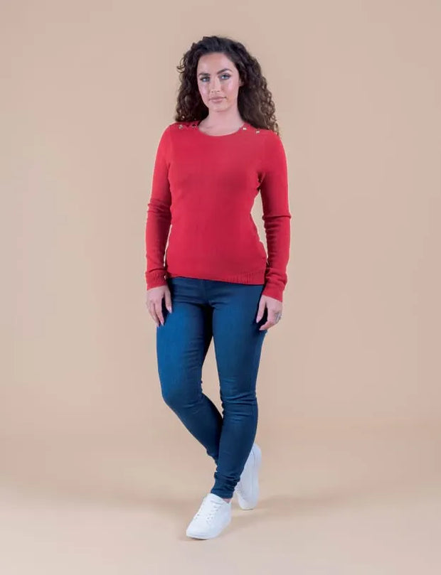 A woman modeling a red long-sleeve knit sweater with shoulder details, paired with dark blue skinny jeans and white sneakers, against a solid beige studio background.