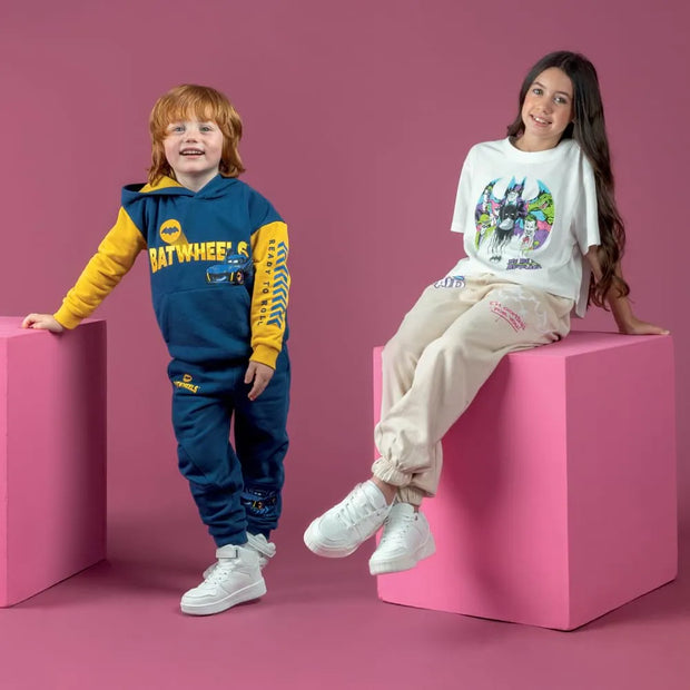 A young boy and a girl modeling Batman-themed clothing against a solid pink studio background. The boy wears a navy blue and yellow Batcrew hoodie and matching pants, while the girl wears a white graphic t-shirt and beige joggers.