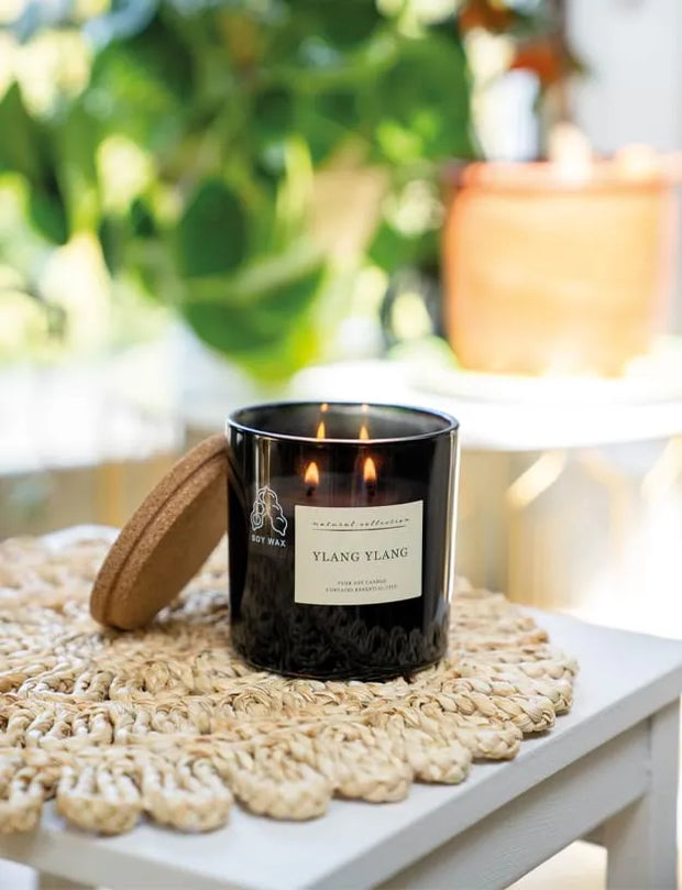 A black glass jar candle with two lit wicks sits on a woven beige placemat on a white table, with a cork lid leaning beside it. The background is softly blurred with green foliage and warm indoor light.