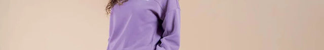 A woman with long curly brown hair is wearing a matching lavender purple sweatshirt and sweatpants set, standing against a solid beige studio background.