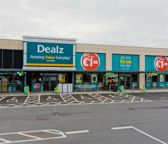 Front exterior view of a retail store under a cloudy sky, featuring a large teal and beige building facade with multiple 'Dealz' signs, red euro coin promotional badges, and an empty paved parking lot in the foreground.