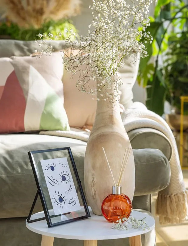 A tall, textured beige ceramic vase filled with delicate white baby's breath flowers, styled on a white side table next to a small amber glass reed diffuser and a black picture frame in a cozy living room setting.