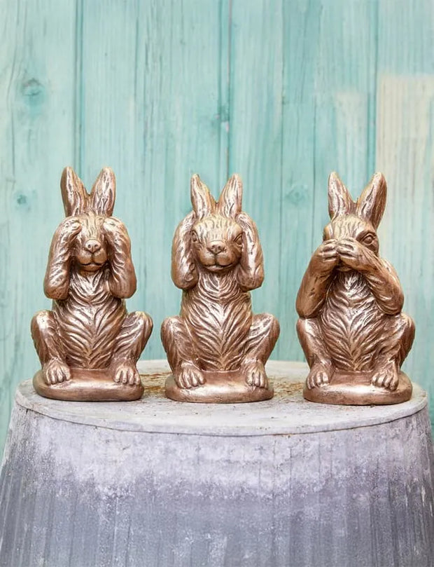 Three bronze-finished rabbit figurines sitting side by side on a round gray pedestal, set against a rustic light blue-green wooden plank background. The left rabbit covers its eyes, the middle rabbit covers its ears, and the right rabbit covers its mouth.