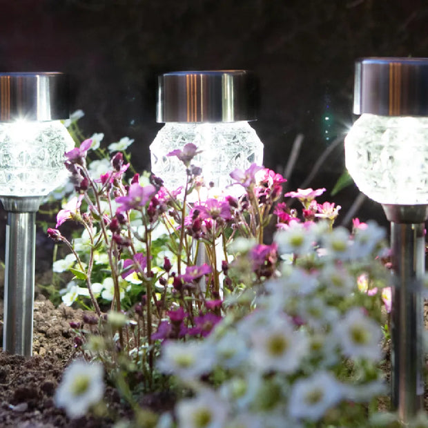 Three solar-powered garden lights with faceted glass shades and metallic caps, glowing in a garden bed with white and pink flowers.