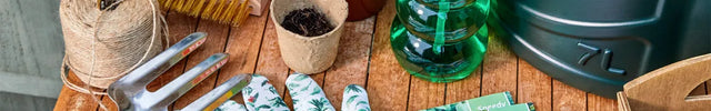 A styled flat-lay of gardening tools and supplies on a wooden table, including a green watering can, green spray bottle, gardening gloves, hand fork, seed packets, twine, and small pots.