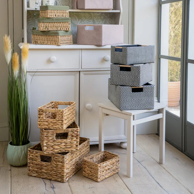 Natural woven storage baskets and fabric storage bins arranged on a floor and stool in a bright, neutral room with a white cabinet and window.