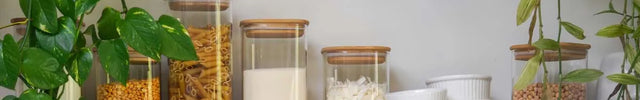 Clear glass canisters with wooden lids filled with pantry staples like pasta, rice, beans, and flour, arranged on a wooden shelf with white bowls and surrounded by green trailing plants against a white wall.
