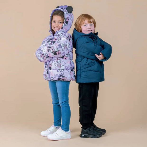 Two children modeling winter jackets against a solid beige studio background. The girl on the left wears a light purple hooded puffer jacket with a cartoon pattern, blue jeans, and white sneakers.