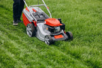 A cordless electric lawnmower with a silver body and orange engine cover being pushed on a green grass lawn. The handle is held by a person whose legs are visible on the left side.