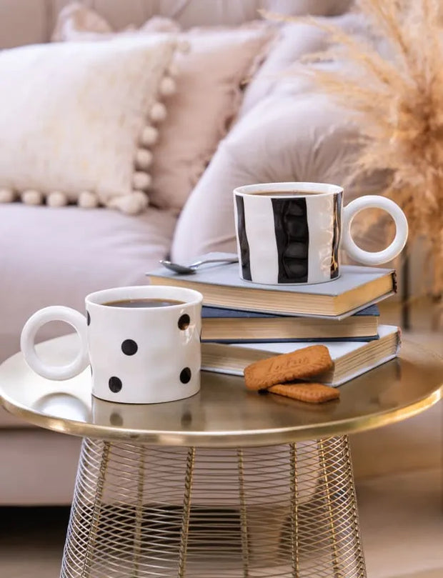 Two ceramic coffee mugs, one white with black polka dots and one white with black vertical stripes, displayed on a stack of books on a round gold wire side table in a cozy living room setting.