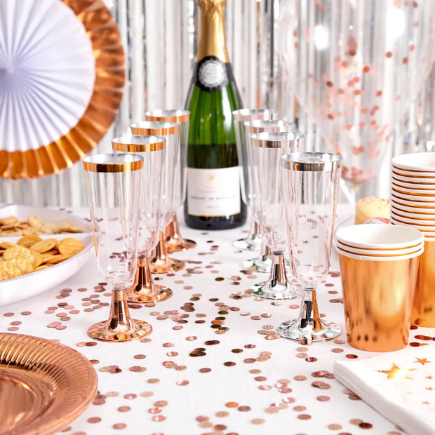Rose gold and clear party tableware set including champagne flutes, paper cups, plates, napkins, and a green champagne bottle on a decorated table with confetti and silver tinsel.