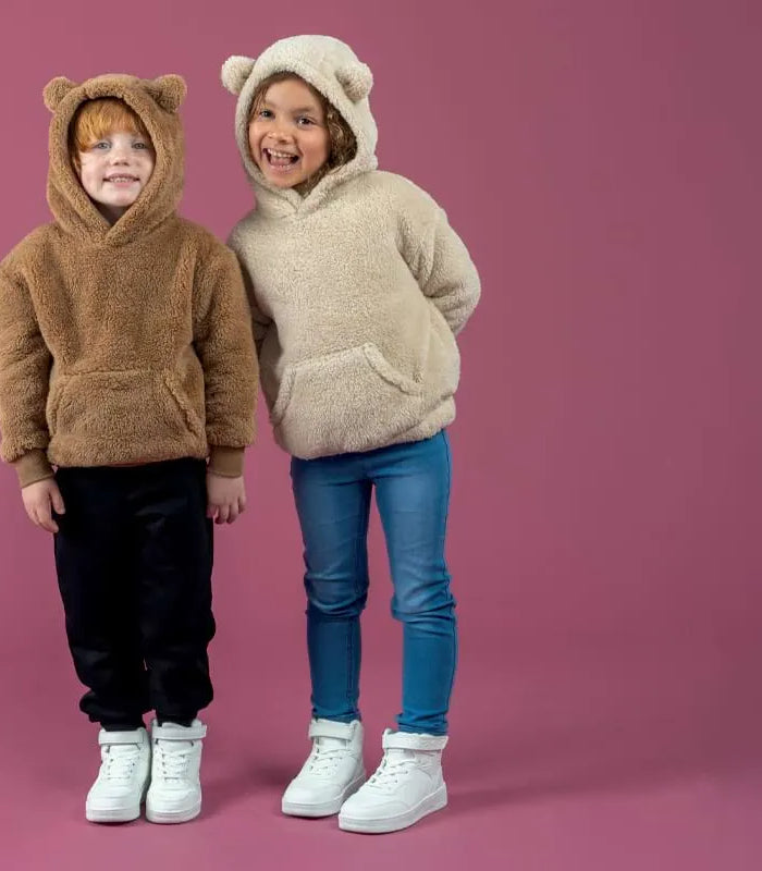 Two children standing side by side in plush teddy-bear hoodies against a solid dusty pink studio background. The child on the left wears a brown hoodie with black pants and white sneakers, and the child on the right wears a beige/cream hoodie with blue jeans and white sneakers.