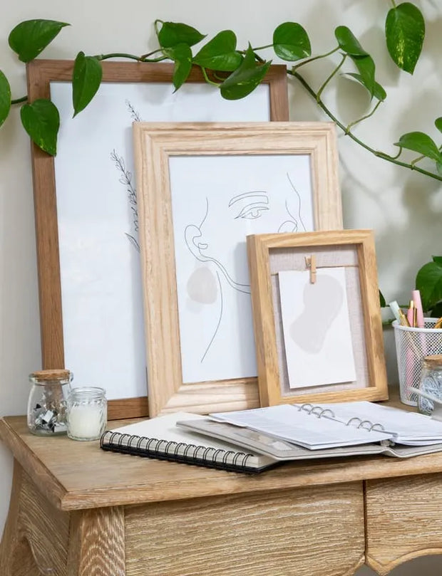 Two light wood picture frames displaying minimalist line art prints, styled on a wooden desk with notebooks, a candle, and a green trailing plant against a white wall.