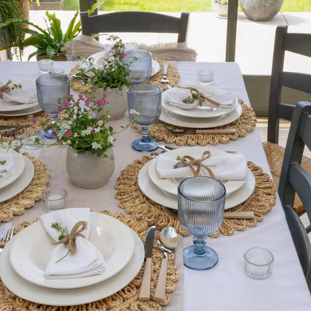 An outdoor dining table set with white plates, white napkins tied with twine, woven round placemats, blue ribbed glass goblets, and small potted flowers on a white tablecloth.