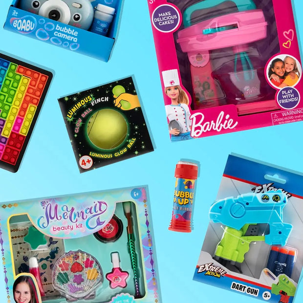 A flat-lay arrangement of various colorful toy products on a light blue background, including a Barbie bubble machine, a Mermaid beauty kit, a pop-it toy, a bubble pop tube, and a dart gun.