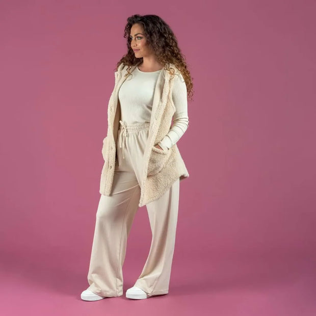 A woman modeling a matching cream loungewear set consisting of a long-sleeve knit top, drawstring waist pants, and white slip-on shoes, styled with an open beige sherpa zip vest against a solid pink studio background.