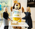 A dog mascot wearing a white t-shirt with 'Dealz Proudly supporting Barretstown' and yellow trousers cuts a cake with two women on a white table in a bright indoor setting.